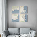 Four square canvas prints featuring vintage-style map designs in muted blue, cream, and beige tones, arranged in a 2x2 grid on a gray wall above a light gray sofa.