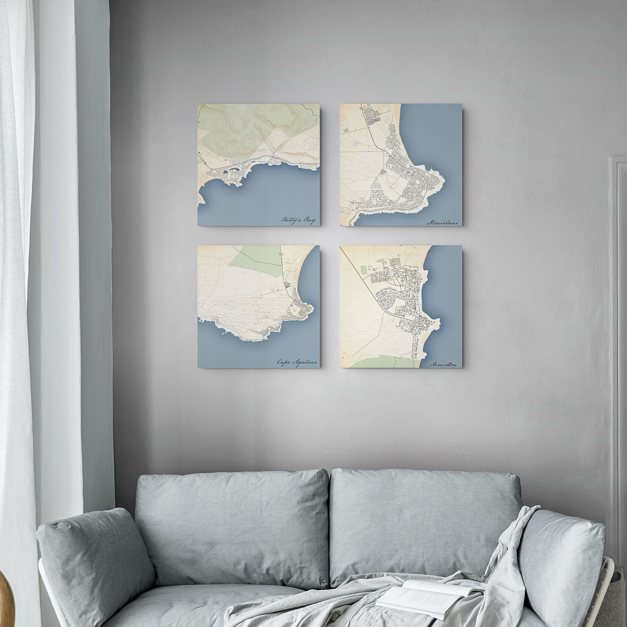 Four square canvas prints featuring vintage-style map designs in muted blue, cream, and beige tones, arranged in a 2x2 grid on a gray wall above a light gray sofa.