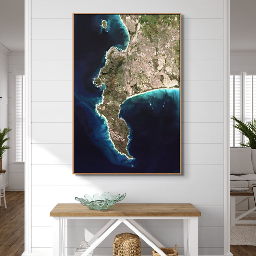 A framed vertical wall art print of the Cape Peninsula in deep blue ocean displayed on a white shiplap wall above a wooden console table in a bright living space.
