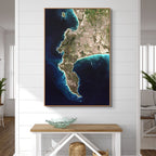 A framed vertical wall art print of the Cape Peninsula in deep blue ocean displayed on a white shiplap wall above a wooden console table in a bright living space.