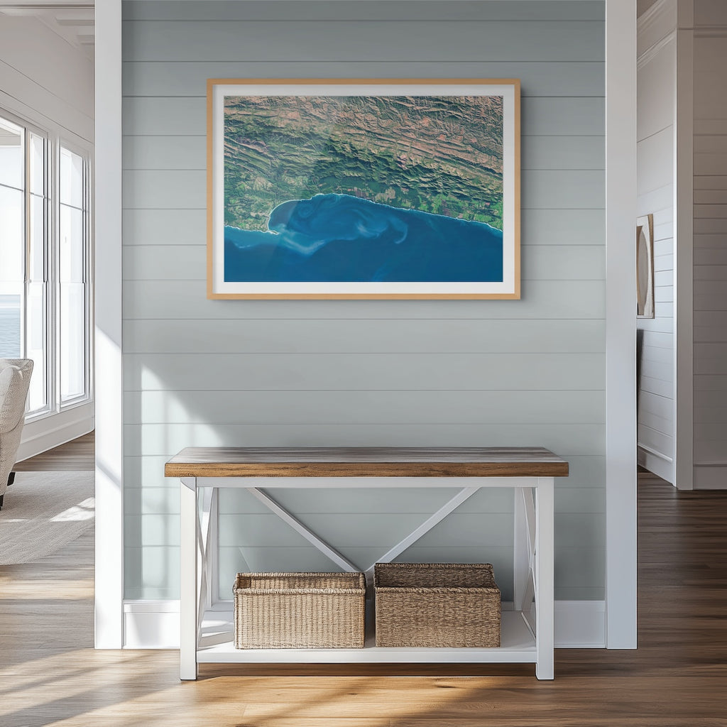 Garden Route 2 East satellite print in a light oak frame above a console table, showing deep blue water and ocean currents along the green coastal mountains.