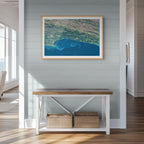 Garden Route 2 East satellite print in a light oak frame above a console table, showing deep blue water and ocean currents along the green coastal mountains.