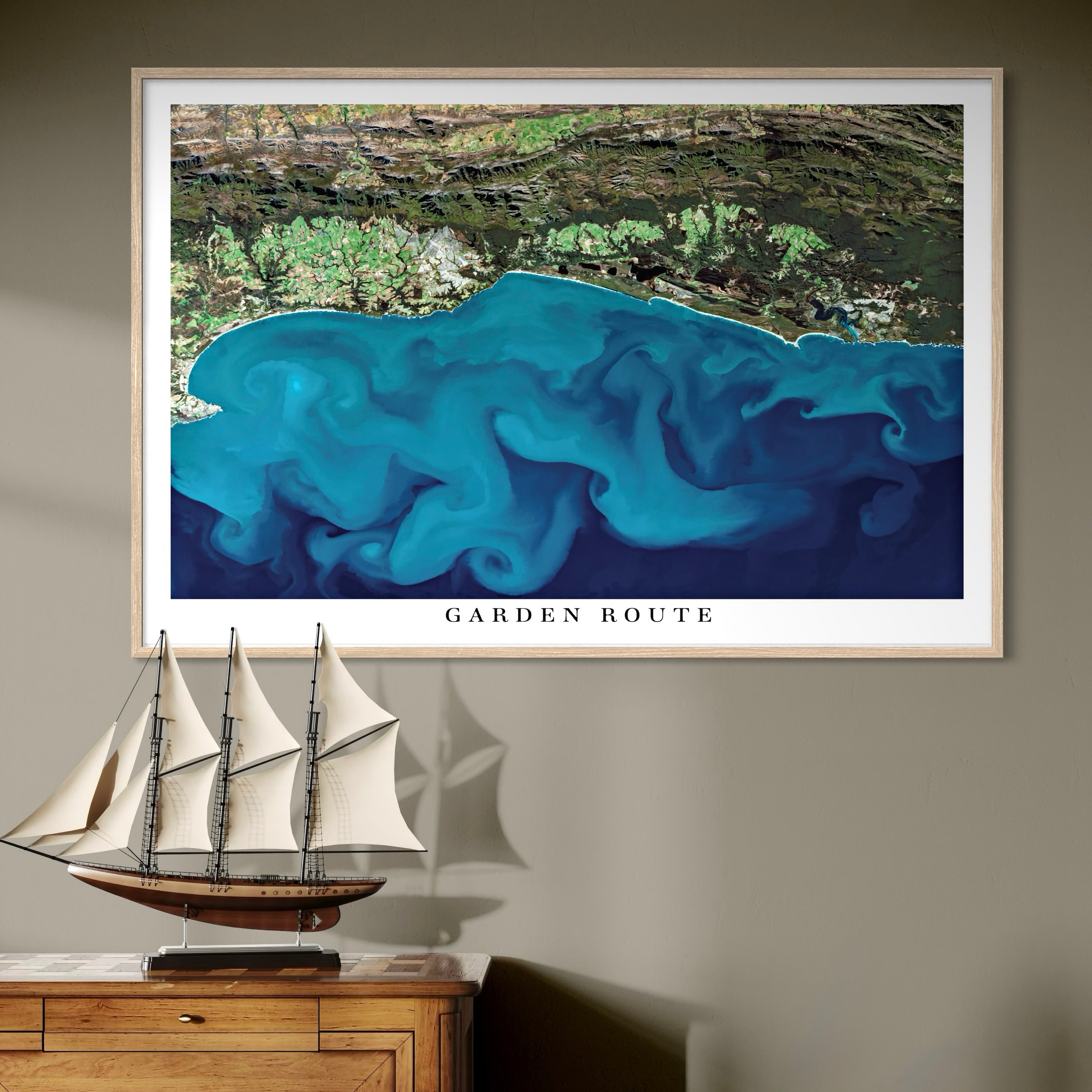 Framed artwork of a blue satellite image titled 'Garden Route' on a wall above a wooden cabinet with a ship model.